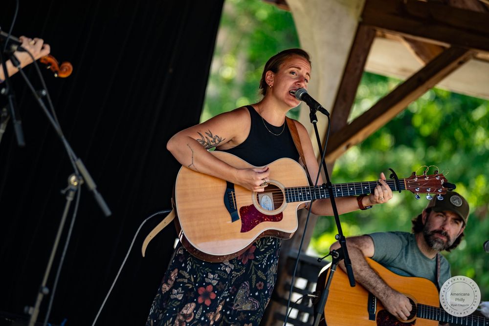 Woman playing acoustic guitar and singing on outdoor stage with male guitarist performing behind her.