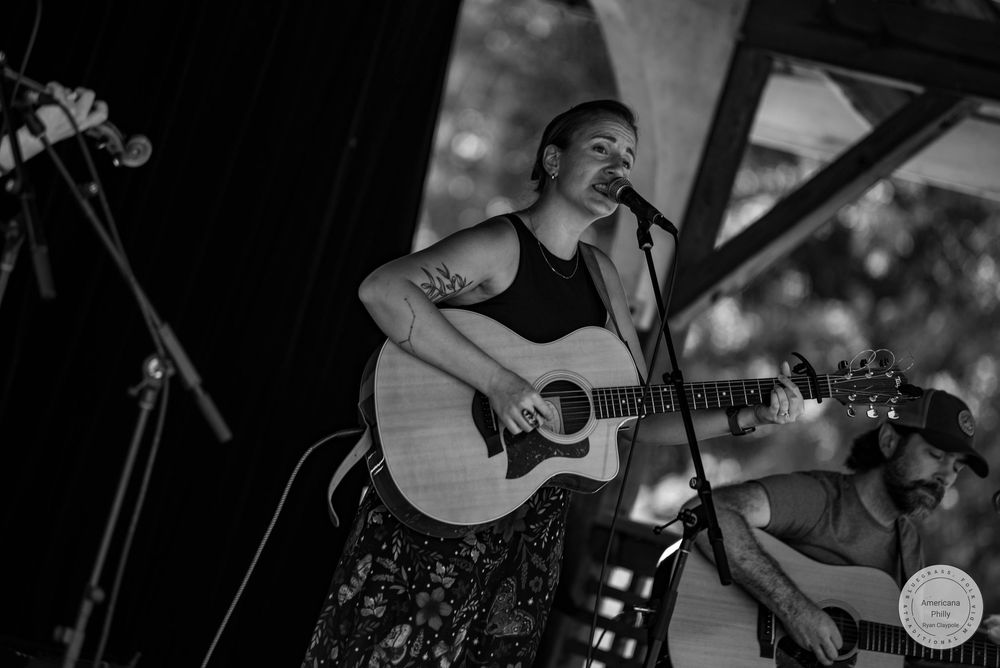 Black and white photo of a woman singing and playing acoustic guitar on stage with another guitarist visible behind her.