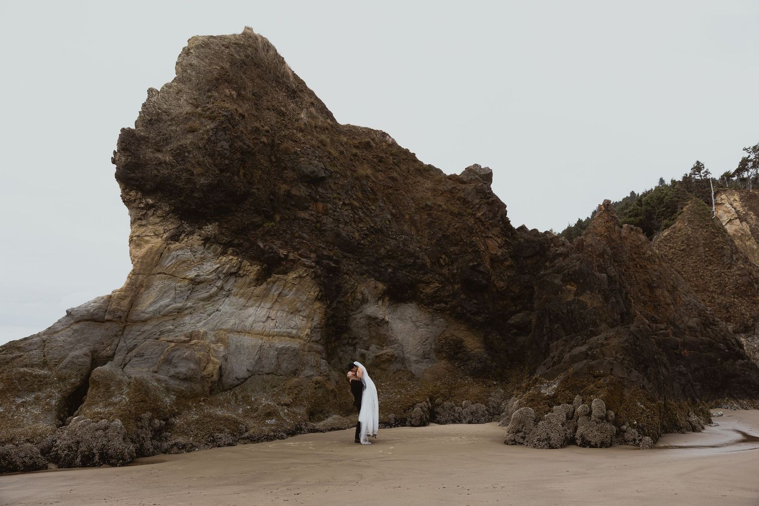 Couple in white attire standing on a sandy beach beside dramatic rocky cliffs on an overcast day.