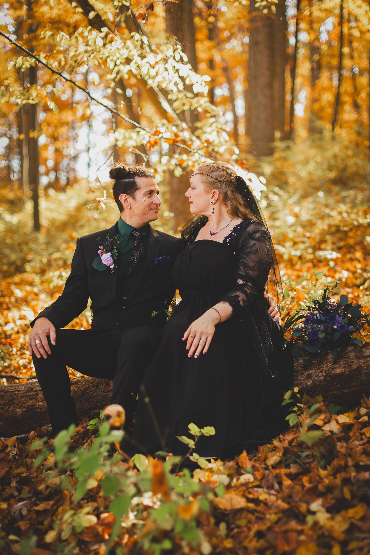 Couple in black wedding attire sitting on a log in an autumn forest with golden leaves at Seneca Creek State Park
