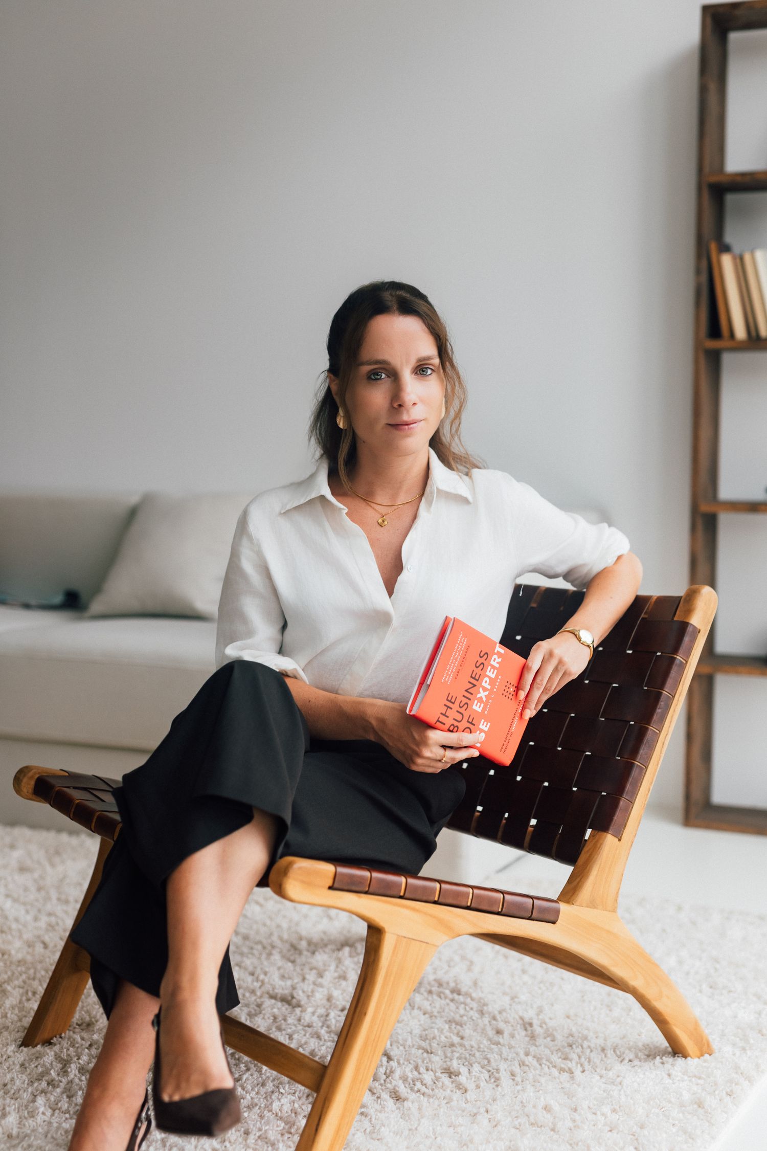 Woman in white blouse holding a red book, seated in a wooden chair in a modern minimalist living room.