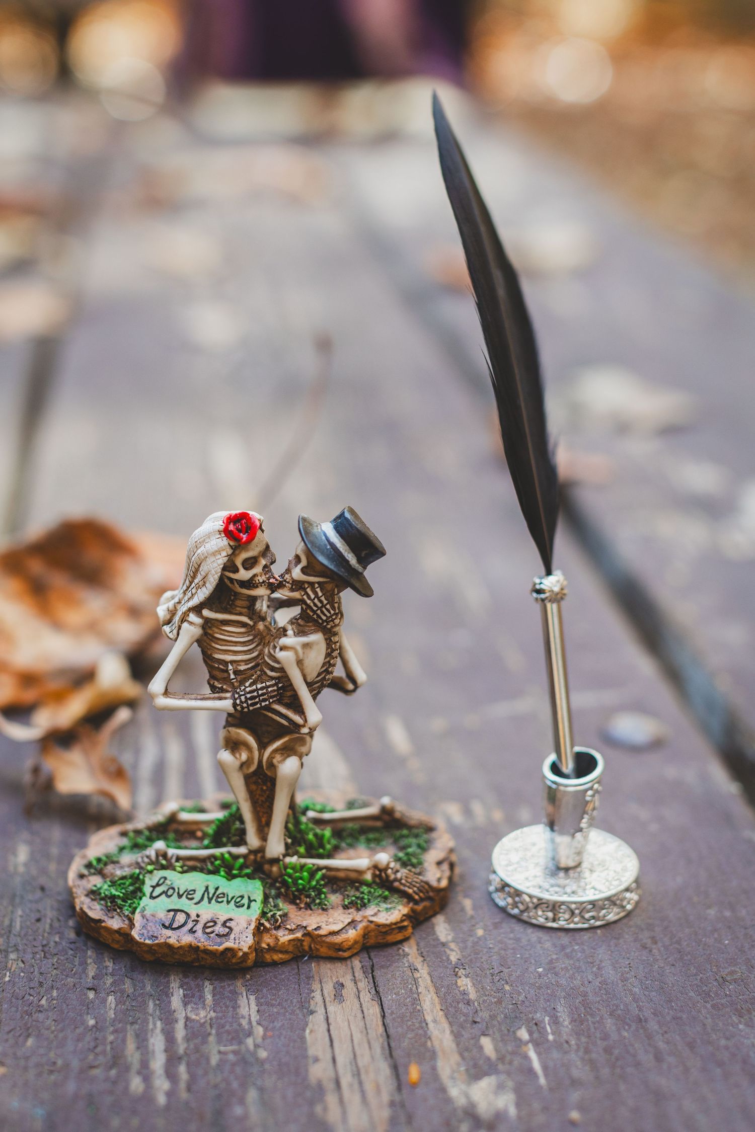 Skeleton couple figurine with Love Never Dies inscription beside a quill pen on a wooden surface.