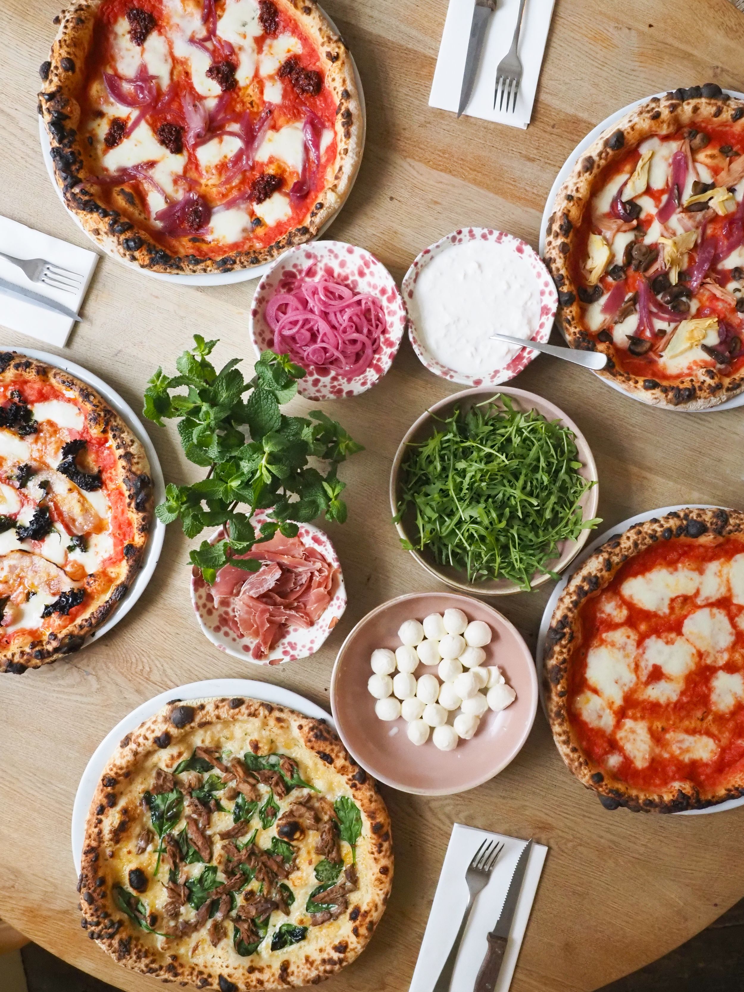 Overhead view of various artisanal pizzas with toppings and garnish bowls on a wooden table.