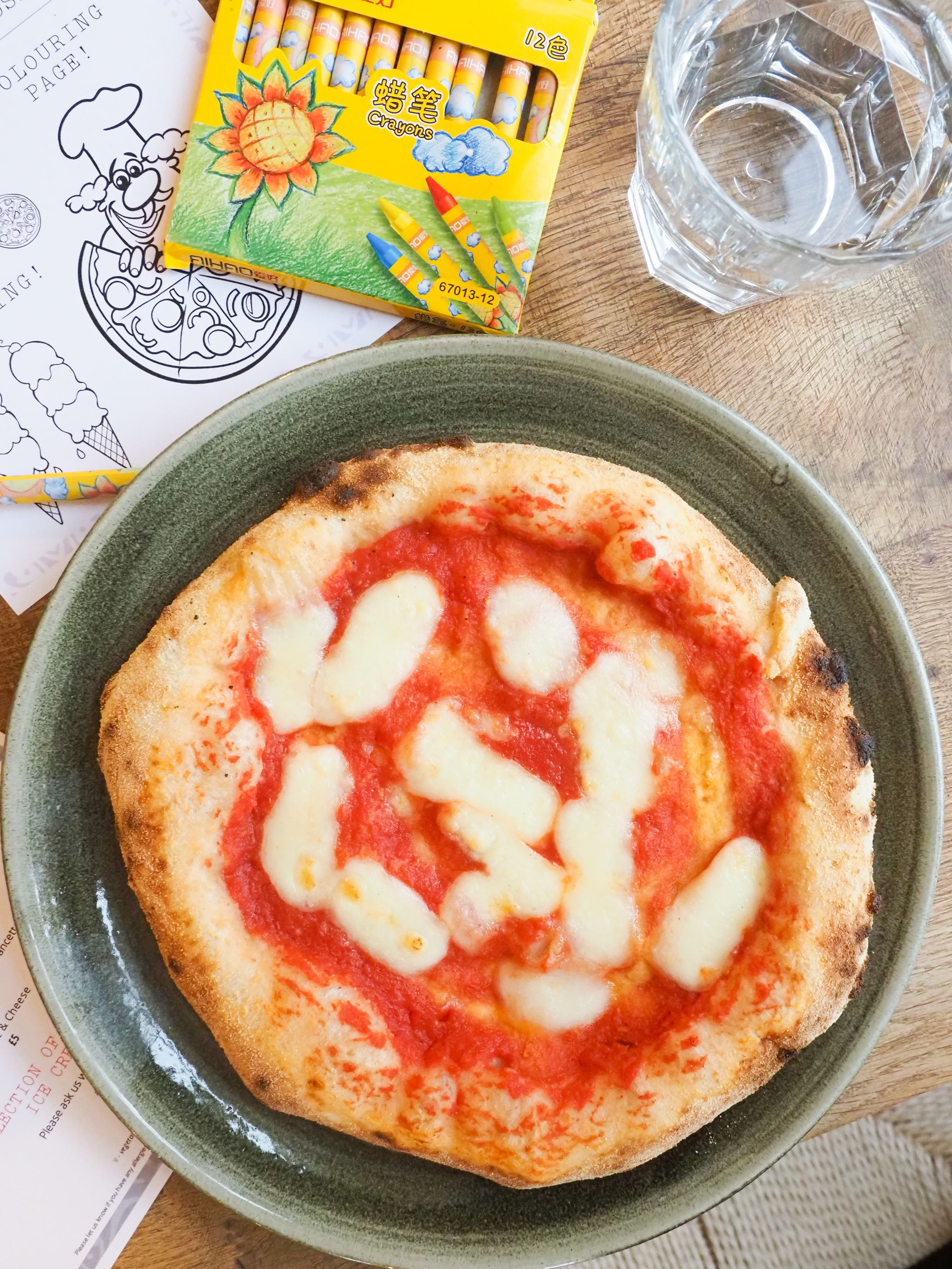 A homemade Margherita pizza with melted mozzarella and red sauce on a gray plate, next to crayons and a glass of water.