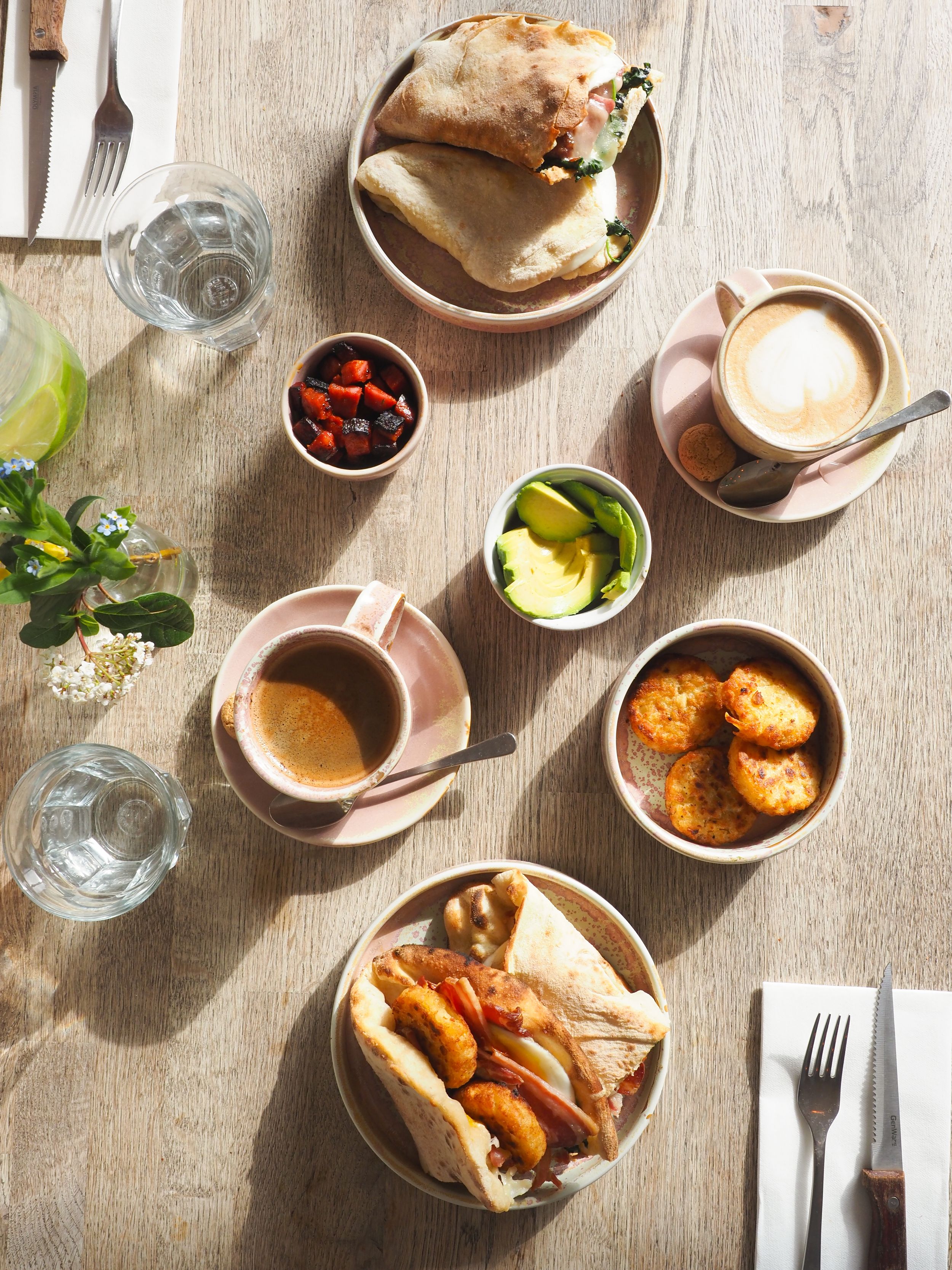 Overhead view of a cafe table setting with coffee cups, small plates of condiments, and sandwiches on a wooden surface.