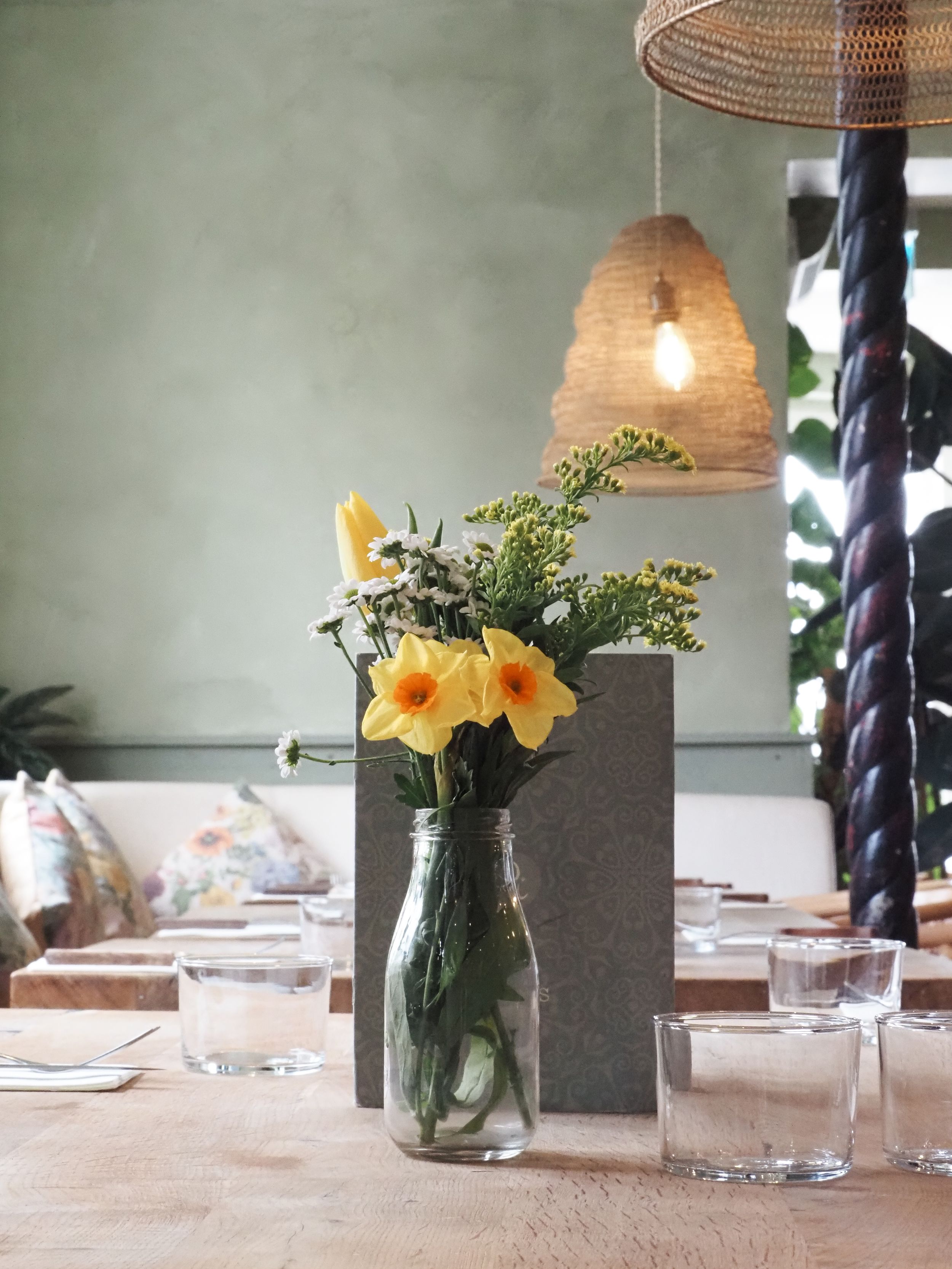 Yellow flowers in glass vase with rattan pendant light above dining table.
