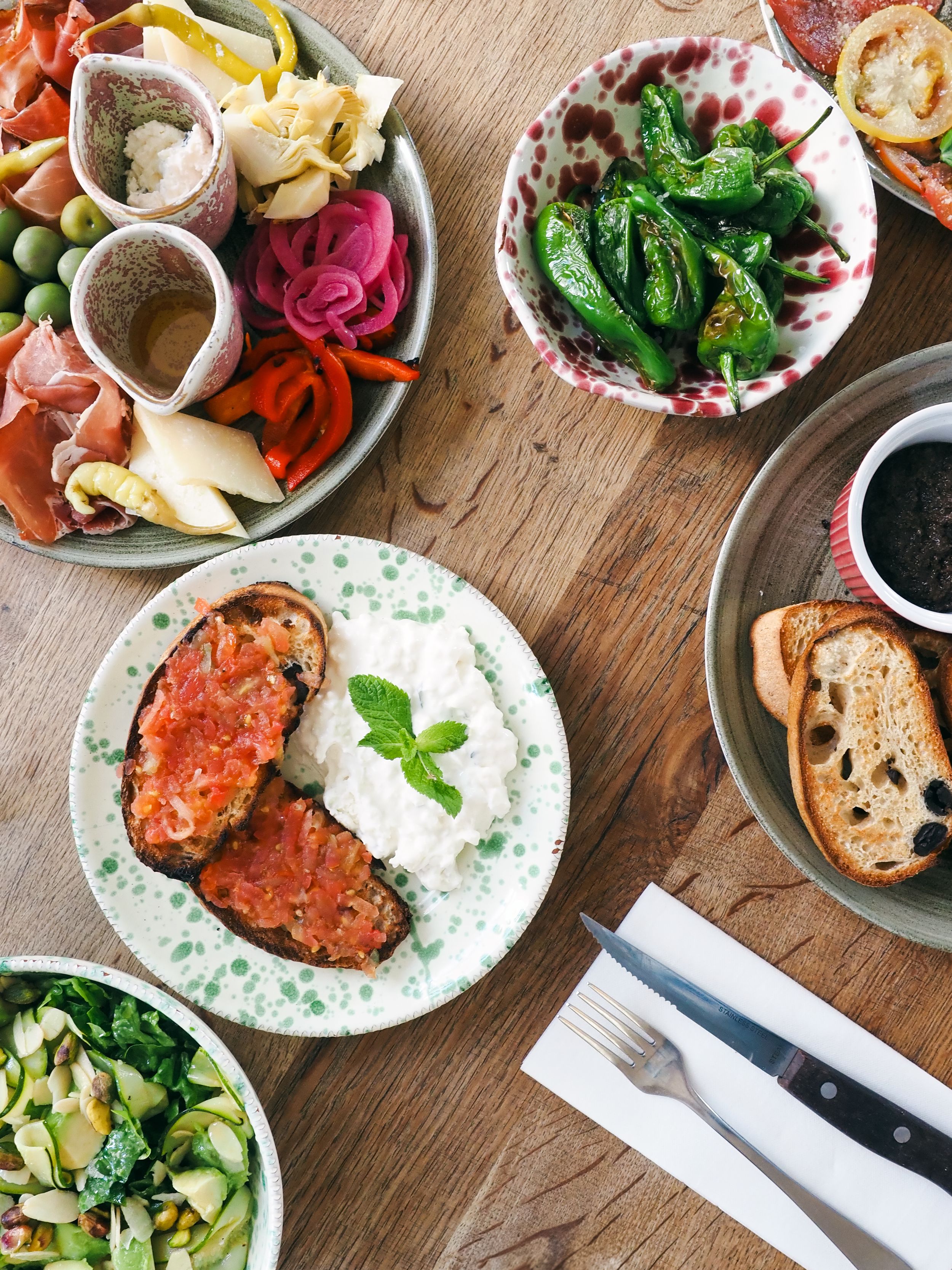 Overhead view of a rustic wooden table set with Mediterranean appetizers including bruschetta, pickled vegetables, and fresh herbs.