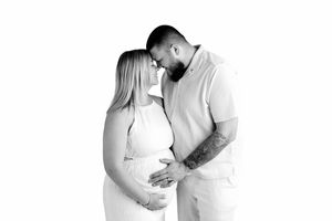 Black and white maternity photo of a couple sharing a tender moment, the woman in a white dress cradling her baby bump.
