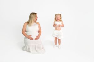 Pregnant woman in white dress sits beside young girl in matching white outfit, both smiling in bright studio setting.