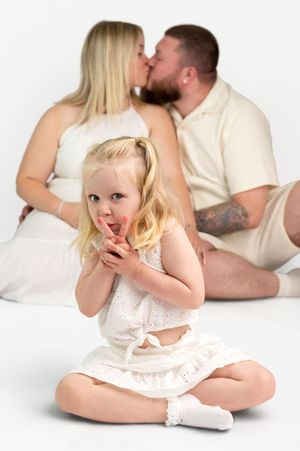 Young blonde girl sits cross-legged in white dress while parents kiss behind her in a bright studio photo.