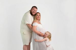 Pregnant woman in white dress poses with partner and young daughter for a family maternity photo shoot.
