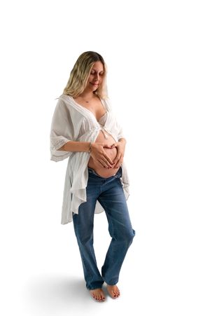 Pregnant woman in white kimono top and jeans forming heart shape with hands on bare baby bump, white background.