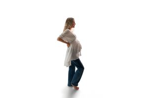 Pregnant woman in white top and blue jeans standing sideways, showing baby bump, barefoot on white background.