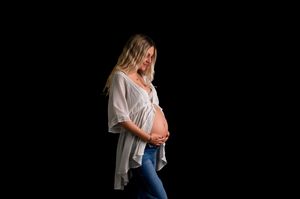 Pregnant woman in white top and jeans cradling her baby bump against a black background.