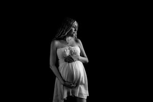 Black and white photo of a pregnant woman in a white dress cradling her baby bump against a dark background.