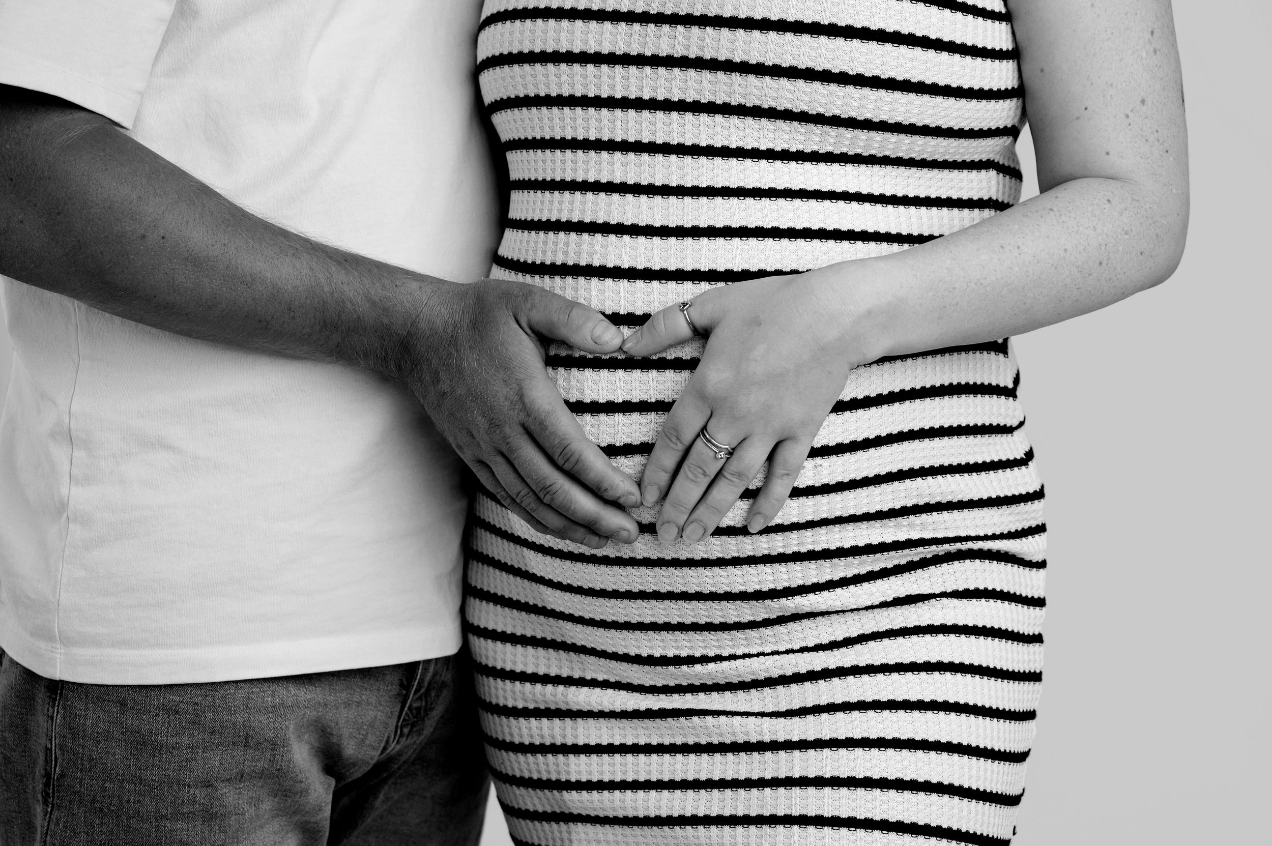 Black and white photo of pregnant couple forming heart shape with hands on baby bump in striped maternity dress.