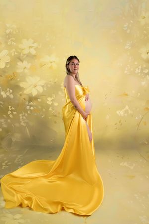 Pregnant woman in elegant yellow satin gown posing against a soft floral yellow background in a maternity portrait.