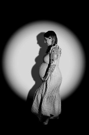 Black and white maternity photo of pregnant woman in gingham dress, illuminated by dramatic spotlight against dark background.
