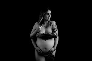 Black and white maternity photo of tattooed pregnant woman cradling bare belly against dark background.