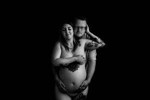 Black and white maternity photo of a tattooed couple embracing, showcasing a pregnant belly against a dark background.