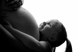 Black and white photo of a young girl hugging a pregnant woman's belly, smiling with joy.