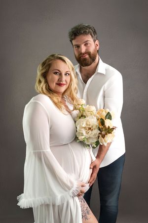 Pregnant woman in white dress holds flowers with bearded man in white shirt during maternity photo shoot.
