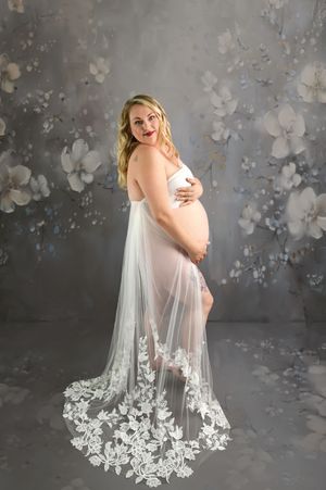 Pregnant woman in white lace maternity gown poses against floral gray backdrop, cradling baby bump.