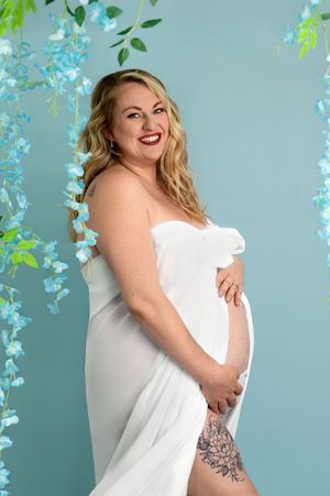 Pregnant woman in white draped dress smiling, holding baby bump, surrounded by blue floral backdrop in maternity photo.