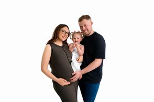 Pregnant woman in gray dress with husband and baby posing for a family maternity photo on white background.