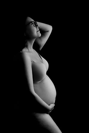 Black and white maternity photo of a pregnant woman in a bra cradling her belly against a dark background.