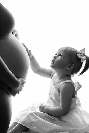 Black and white photo of a toddler girl in white dress touching pregnant mothers belly, looking up with curiosity.