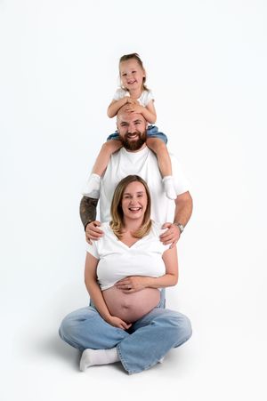 Happy pregnant family with bearded dad, young daughter on shoulders, and expectant mother sitting cross-legged on white background.