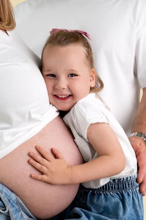 A smiling young girl hugs her mother's pregnant belly while sitting together on a white bed.