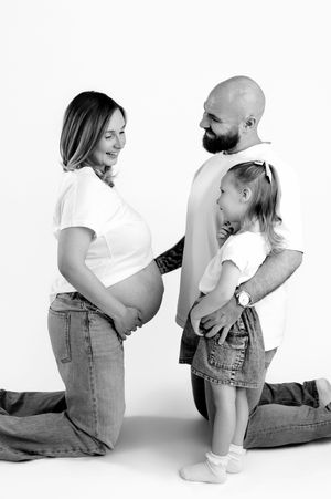 Black and white maternity photo of pregnant woman, bald bearded man, and young girl posing together on white background.