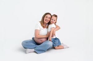 Pregnant woman in white tee and jeans sits cross-legged with toddler on white background.