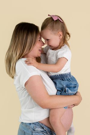 Pregnant mother in white tee holds toddler girl in denim skirt, both smiling and touching foreheads tenderly.