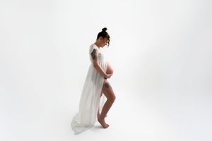 Pregnant woman in flowing white sheer gown poses sideways showing baby bump against white background.
