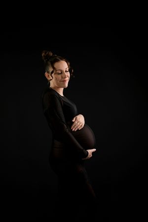Pregnant woman in black outfit holding baby bump, elegant side profile maternity photo on dark background.