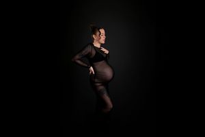 Pregnant woman in black sheer dress posing sideways against dark background in elegant maternity photo.