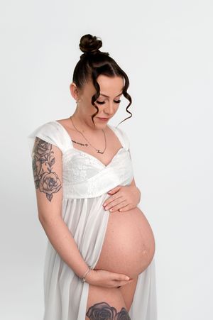 Pregnant woman in white lace dress cradling baby bump, featuring floral tattoos, studio photo on white background.