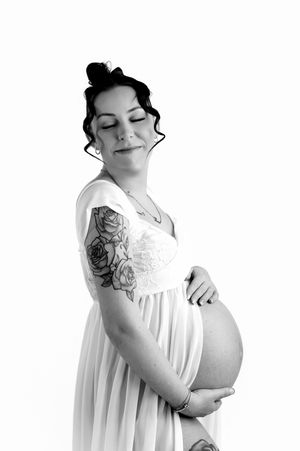 Black and white maternity photo of a tattooed pregnant woman in a white dress cradling her baby bump with eyes closed.