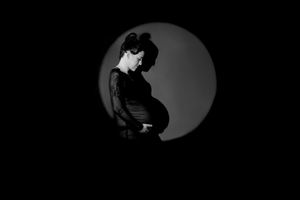 Black and white maternity photo of pregnant woman in lace dress, posed in circular spotlight against dark background.