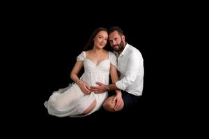 Expecting couple in maternity photo shoot, woman in white dress, man in white shirt, black background.