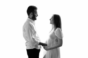 Black and white photo of a smiling couple holding hands, pregnant woman in white dress facing man in white shirt.