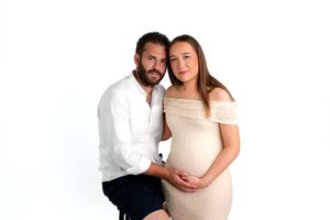 Couple posing for maternity photo shoot, pregnant woman in beige dress with partner in white shirt on white background.