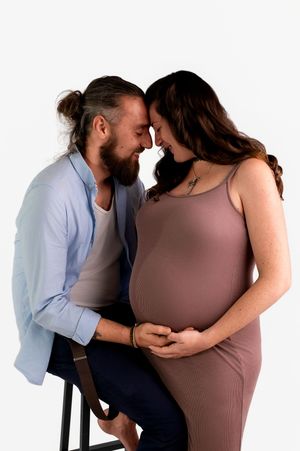 A smiling couple sharing a tender forehead touch, with the pregnant woman in a mauve dress cradling her baby bump.