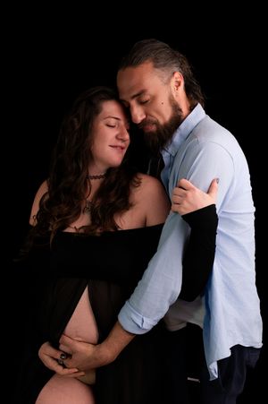 Couple sharing tender maternity photo moment, pregnant woman in black dress embraced by bearded man, black background.