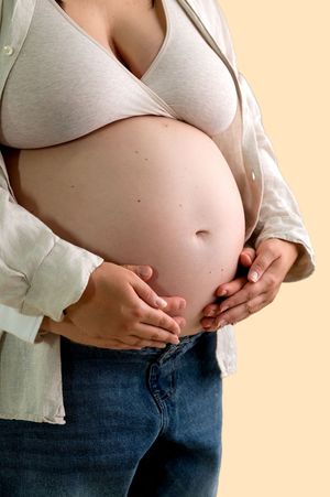 Pregnant woman in open white shirt and jeans cradling her bare baby bump with both hands on beige background.