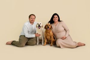 Couple sitting on floor with two dogs, yellow Lab and golden retriever, pregnant woman in beige dress, neutral background.