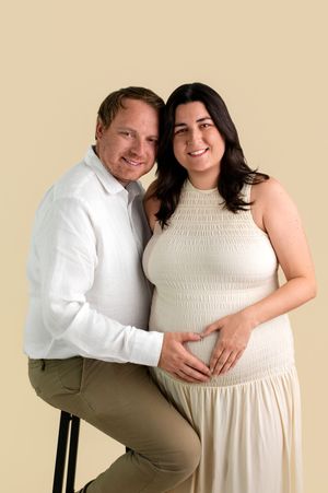 Happy couple posing for maternity photo, woman in cream dress cradling baby bump, man in white shirt, beige background.